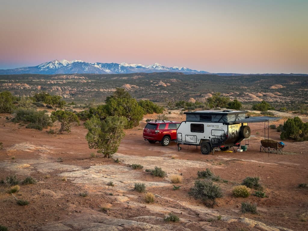 An overland camper and SUV camped in the desert at sunset with snow capped mountains in the background.