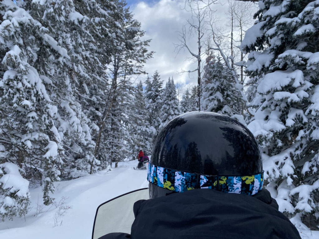 Up close shot of a snowmobiler with a black helmet going though a snow covered pine forest. 