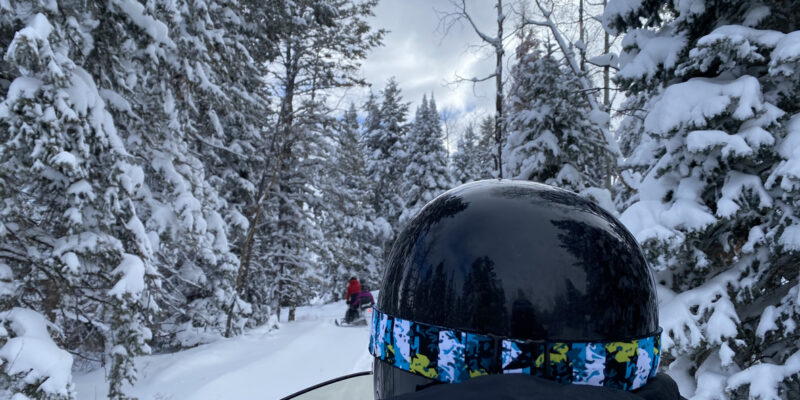 Up close shot of a snowmobiler with a black helmet going though a snow covered pine forest.