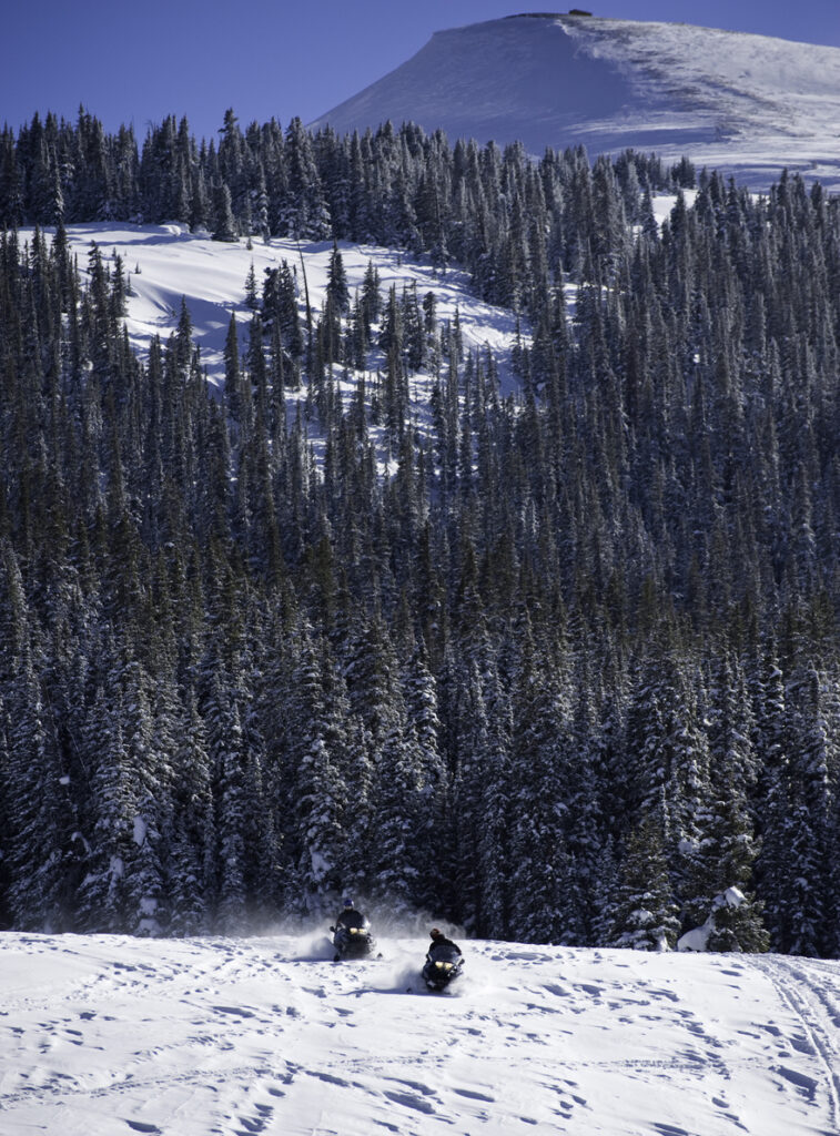 Two snowmobilers going down the side of a snowy mountain in Colorado as they enjoy snowmobiling in Colorado. 