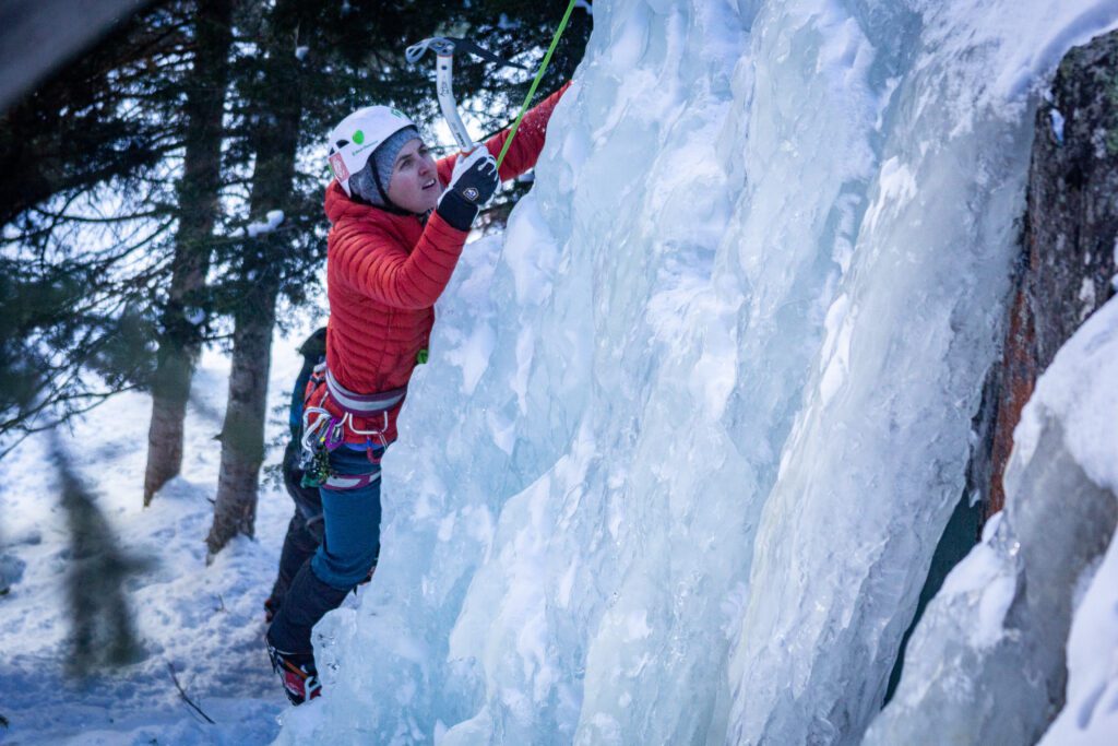 Meg in a red jacket scaling a wall of ice with a helmet on during winter in Colorado.