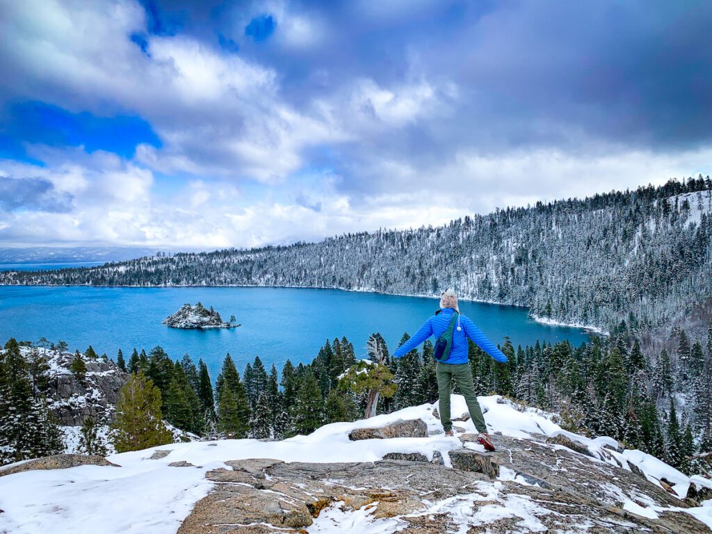 A person in a blue jacket and white hat looking out at a stunning view of Lake Tahoe with clear skies and snow on the ground in January in Lake Tahoe.