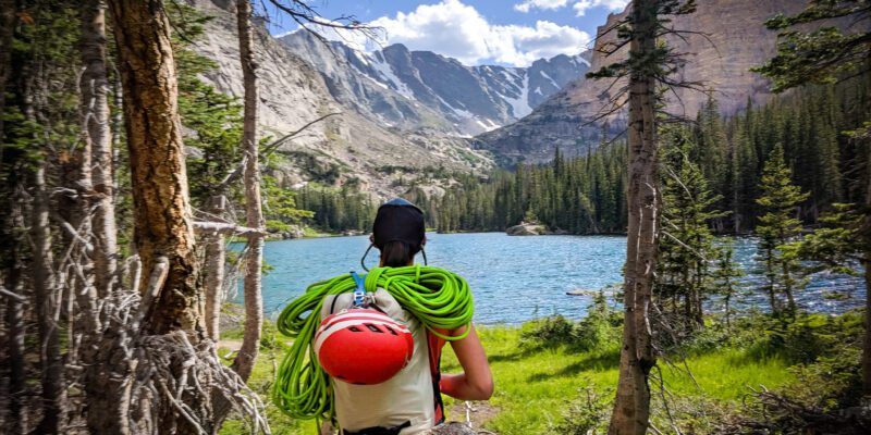 Meg is staring at an alpine lake in RMNP. She has a backpack on with green rope and a red helmet.