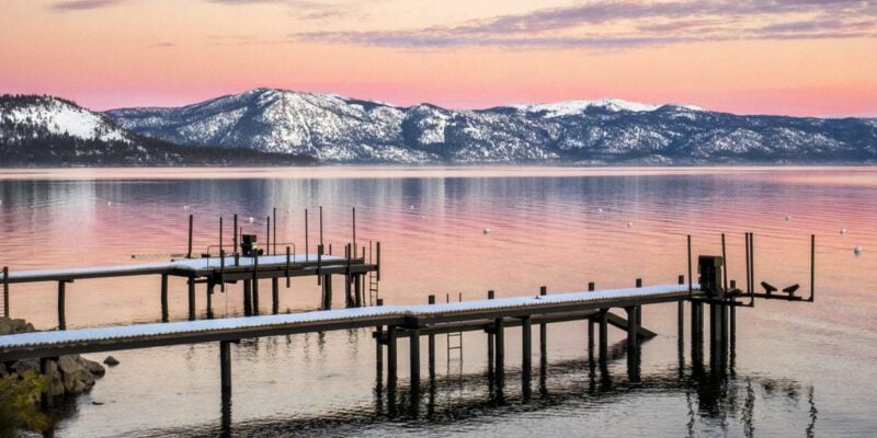 A view of two docks covered in snow in Lake Tahoe in February. The sun is setting in the background and the mountains are covered in snow.