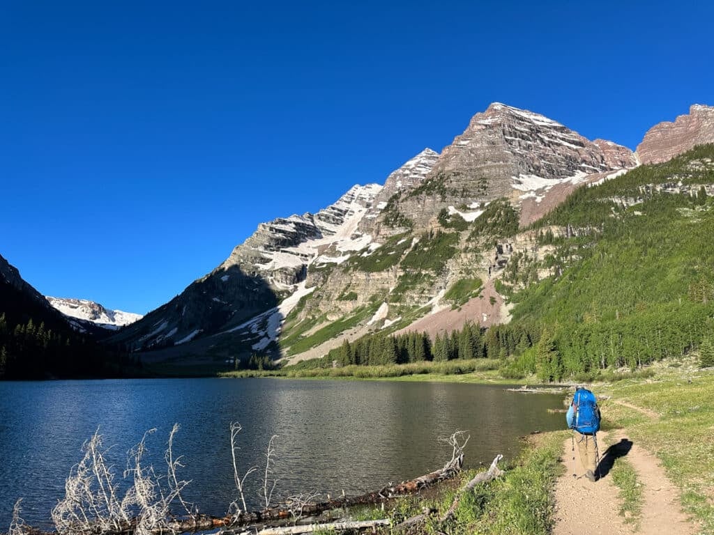 A man walking down a trail next to tall mountains dusted in snow with a lake on their side.