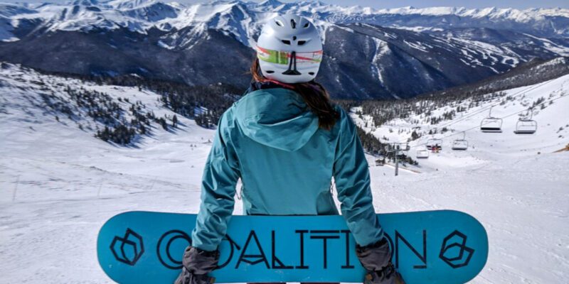 A woman is holding a blue snowboard behind her back and has a turquoise jacket on. She has long dark hair, is wearing a white helmet, and is looking at the snow covered mountains and slopes of Colorado.