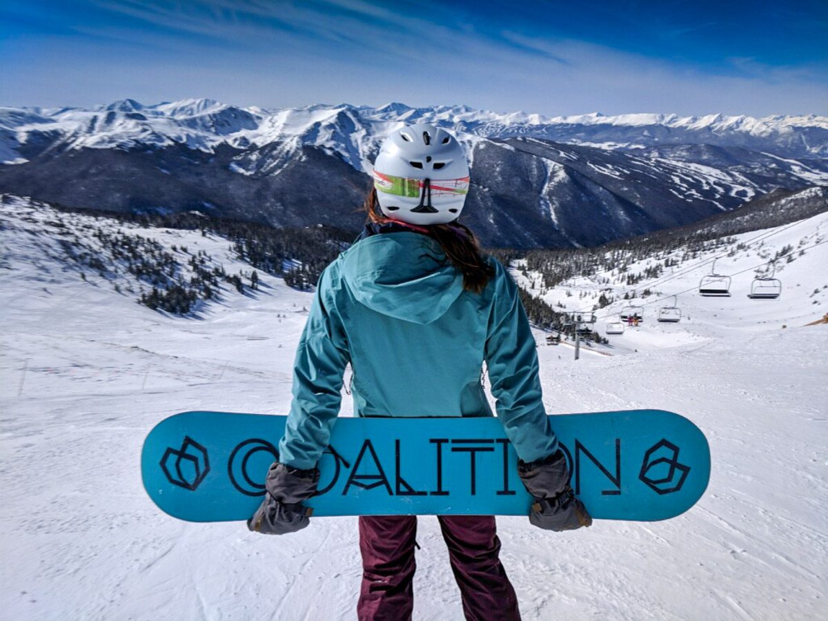A woman is holding a blue snowboard behind her back and has a turquoise jacket on. She has long dark hair, is wearing a white helmet, and is looking at the snow covered mountains and slopes of Colorado.