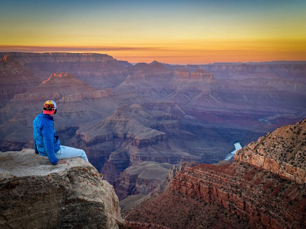 Person sitting on the edge of the Grand Canyon wearing jeans, a long-sleeve blue shirt, and a backwards baseball cap watching the sun set over the Grand Canyon.
