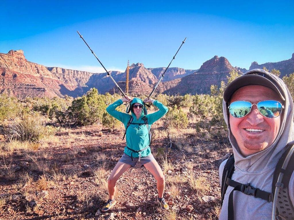 Meg standing in a long, greenish shirt and shorts with hiking poles sticking up and out of her ears. A man is standing with sunglasses close to the camera with a hat and you can see the natural landscape of the Grand Canyon in the background. 