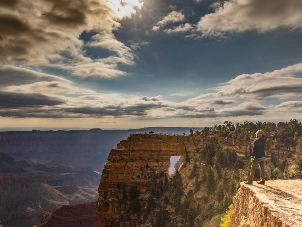 A person is looking out at the North Rim of the Grand Canyon with a stone arch on the edge of the canyon in front of them. 