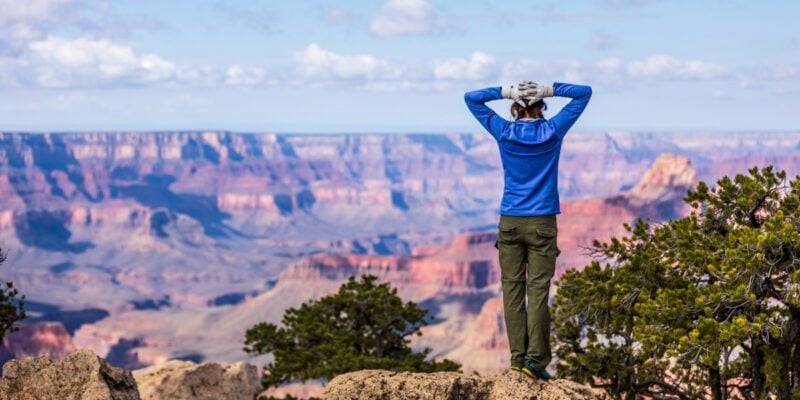 Meg is standing in a long sleeve blue shirt and pants. Her hands are behind her head and she is staring out at the Grand Canyon from one of the best hiking trails in the Grand Canyon.
