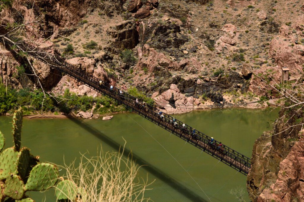 Aerial view of a narrow bridge across a green river in the Grand Canyon. People are walking across a bridge between two mountains that is a designated mule crossing. 