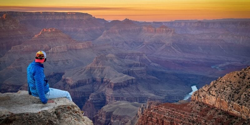 Person wearing a long blue-sleeve t-shirt and a backward cap looking out at the sun setting over the Grand Canyon.