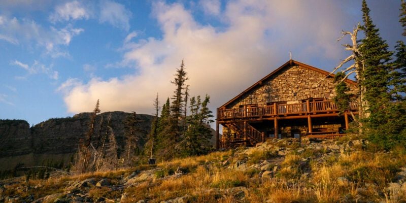 A wooden building sitting on the side of a mountain at sunset in Granite Park. The skies are blue and filled with clouds.