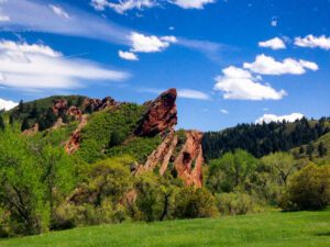 Vibrant red rocks jutting out of the landscape and surrounded by green grass and trees on a bright day with a blue sky with clouds in the sky.