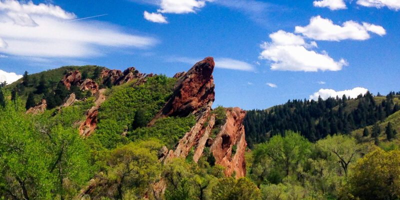 Vibrant red rocks jutting out of the landscape and surrounded by green grass and trees on a bright day with a blue sky with clouds in the sky.