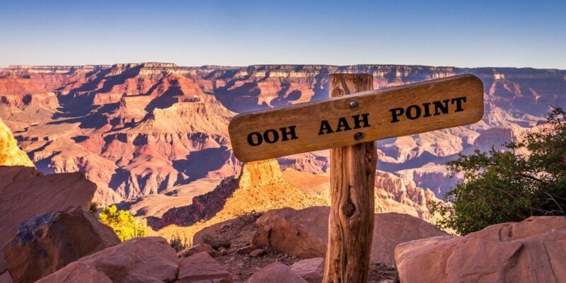 A view of a wooden sign that says, "Ohh Ahh Point" sitting in front of the Grand Canyon during sunrise.