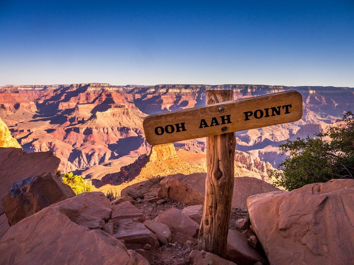 A view of a wooden sign that says, "Ohh Ahh Point" sitting in front of the Grand Canyon during sunrise.