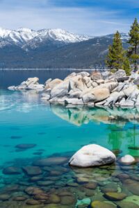 View of Lake Tahoe on a sunny day. You can see the rocky shore and the snow-covered mountains in the background and turquoise water in the foreground.