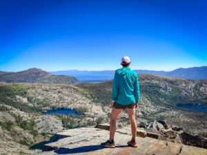 A woman standing on top of Ralston Peak in the Desolation Wilderness on a cloudless day. A deep blue Lake Tahoe is in the background with several smaller lakes in the foreground.