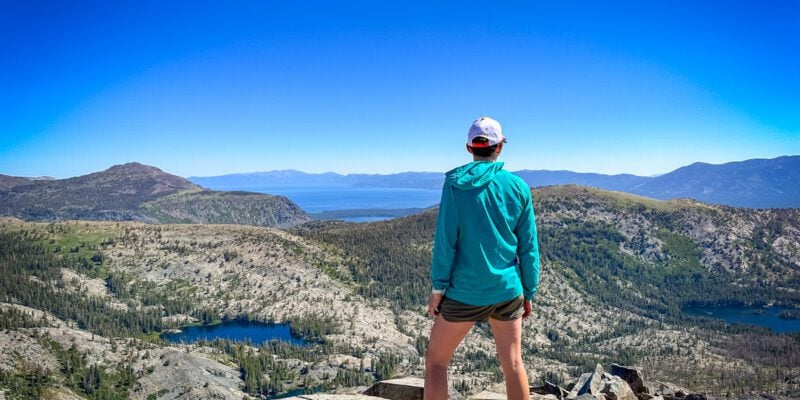A woman standing on top of Ralston Peak in the Desolation Wilderness on a cloudless day. A deep blue Lake Tahoe is in the background with several smaller lakes in the foreground.