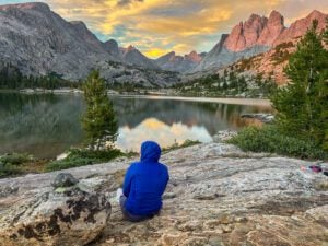 A woman sitting next to a still mountain lake called Bonneville Lake with sharp, dramatic mountain peaks in the distance.