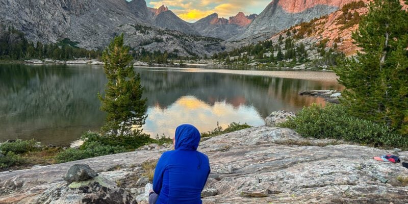 A woman sitting next to a still mountain lake called Bonneville Lake with sharp, dramatic mountain peaks in the distance.