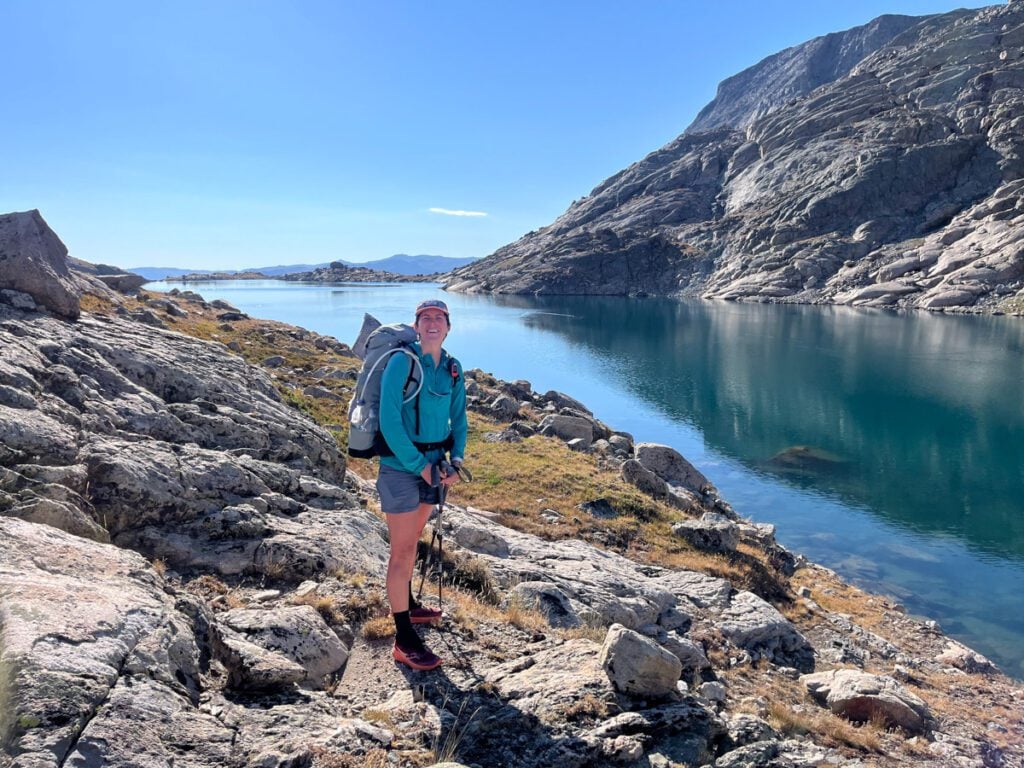 A woman standing by a still alpine lake in the Wind River Range.