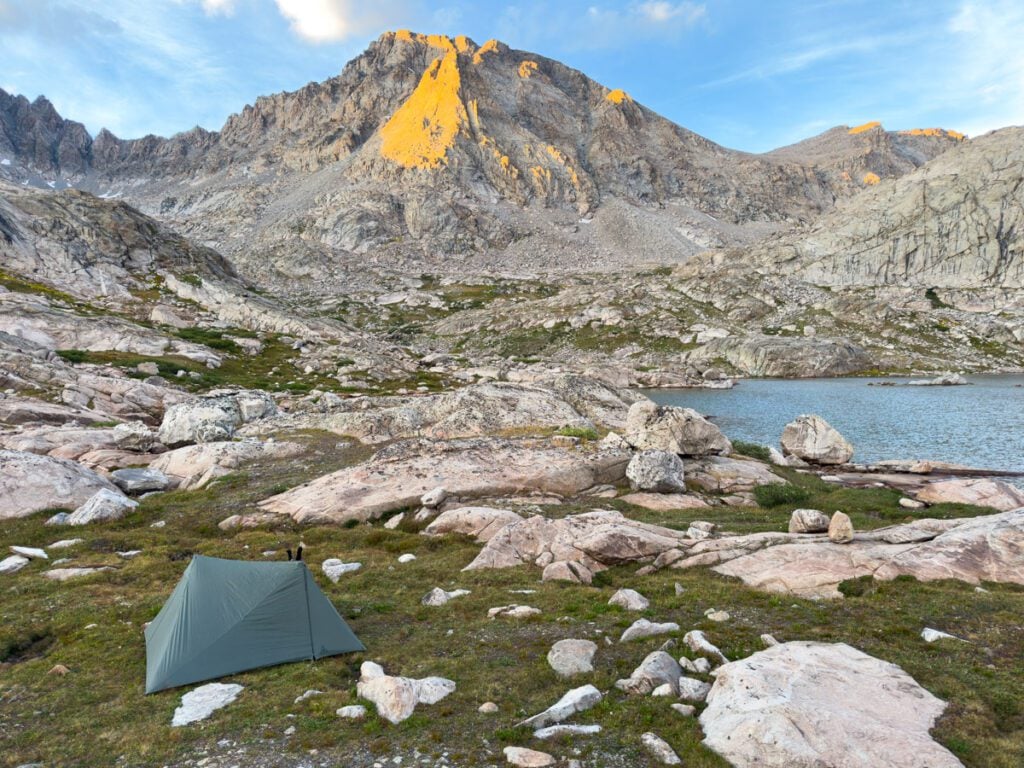 A tent sitting near an alpine lake with a tall, rocky mountain peak in the background in Indian Basin.