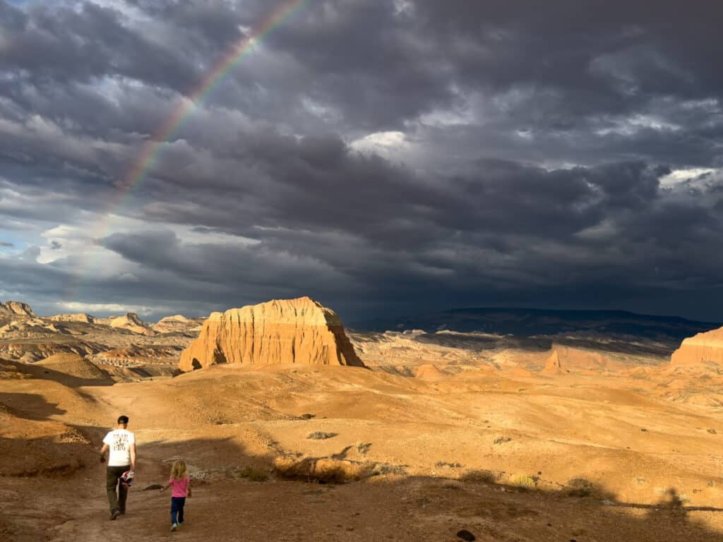 A dad and daughter hiking under a rainbow in Cathedral Valley