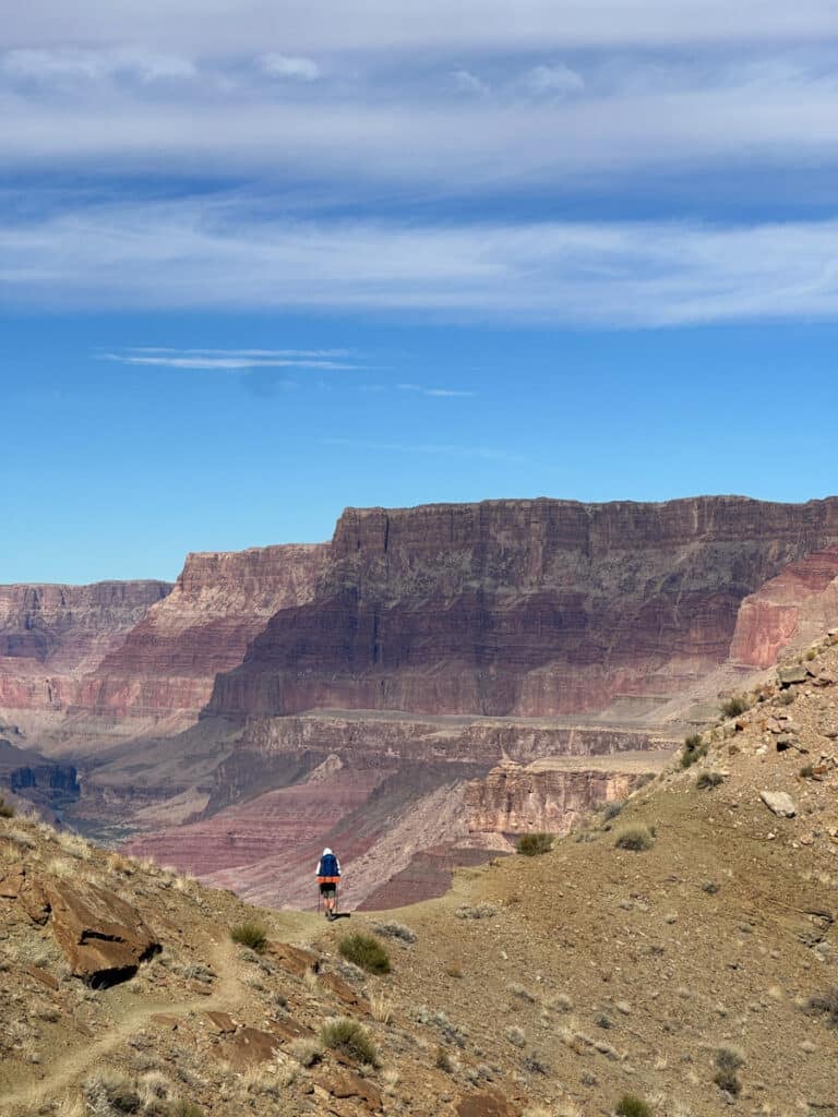 A man standing at a ridge in the grand canyon overlooking the palisade cliffs, steep red wall cliffs that drop down to the mighty colorado on a partly cloudy day.