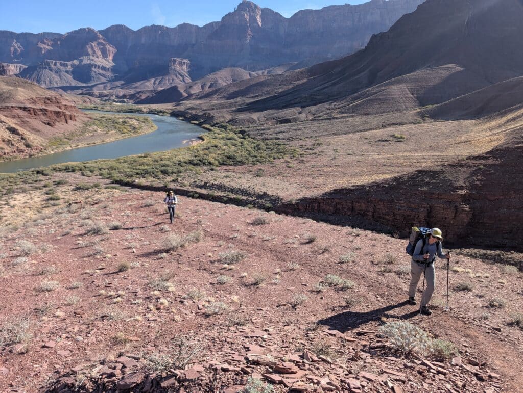two people climbing a big red rock hill in the Grand Canyon with the Colorado River in the background. 